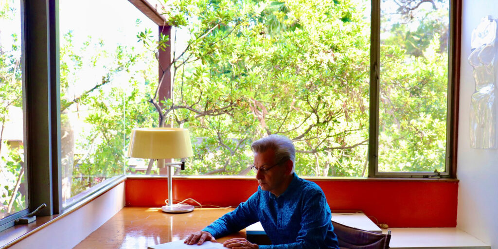 A man sits in a corner office with abundant light. The seamless windows disappear connecting the inhabitant more closely with nature